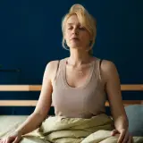 Kvinna med slutna ögon sitter i sängen, insvept i täcke, i en avslappnad somna meditation.