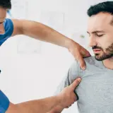 Fizjoterapeuta bada bark pacjenta, który odczuwa ból. Objawy uszkodzenia barku mogą być różne.