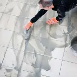 Man med orange handskar arbetar med att måla fogar på ett kaklat golv. En hink med fogmassa står bredvid.
