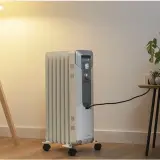 Estufa eléctrica de aceite con ruedas, conectada a la corriente, junto a una planta y una lámpara. Controla el consumo.
