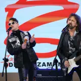 Zespół rockowy na scenie, wokalista w białych okularach i kolorowej kurtce, grający na marakasach. Obok gitarzysta w skórzanej kurtce. W tle logo "Jary Oddział Zamknięty".