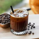 Erfrischender kalter Kaffee mit Eiswürfeln und einem Strohhalm, daneben eine Schale mit Kaffeebohnen.