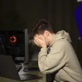 Un joven frustrado cubre su rostro, rodeado de equipos informáticos. Parece que su PC sufre un cuello de botella y no puede jugar.
