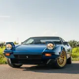Niebieski De Tomaso Pantera z wyskakującymi reflektorami i złotymi felgami na tle zieleni i błękitnego nieba.
