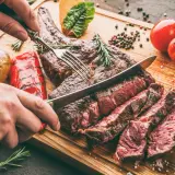 Une main coupe un succulent côte de bœuf barbecue sur une planche en bois, accompagné de pommes de terre rôties et de poivrons grillés.