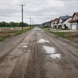 Błotnista droga polna prowadzi do szeregu nowych domów. Po lewej stronie widać niezabudowane działki z tabliczkami.