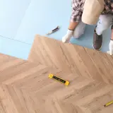 Instalator w białych rękawiczkach układa panele podłogowe w jodełkę. Widać poziomicę i miarkę, co sugeruje, jak kłaść panele.