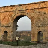 Imponente arco romano de piedra, con dos arcos menores a los lados, se alza sobre un paisaje de colinas verdes.