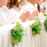 Dzieci w bieli, z modlącymi się dłońmi, czekają na komunię. Dekoracje z winogron i zieleni podkreślają uroczystość, a koszty komunii w kościele są nieodłącznym elementem tego dnia.