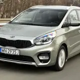 Srebrny Kia Carens 4 w ruchu drogowym, z widocznym grillem i reflektorami.
