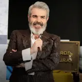 Lorenzo Caprile, con su característica barba canosa, sonríe mientras sostiene unas gafas. Viste una chaqueta de tweed con bordados florales.