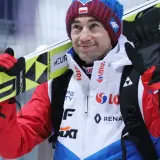 Uśmiechnięty Andrzej Stoch Jr. z nartami na ramieniu, gotowy do skoku.