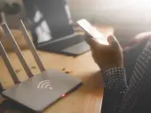 Router Wi-Fi, smartfon w dłoni, laptop na drewnianym biurku.