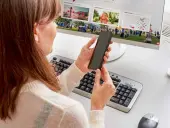 Kobieta trzyma smartfon przed monitorem komputera z klawiaturą i myszką.