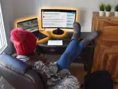 Kobieta w czerwonej czapce i szarych kozakach pracuje przy laptopie i zewnętrznym monitorze.