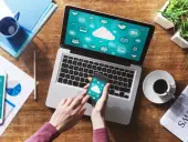 Dłonie trzymające smartfon z ikonami chmury na tle laptopa i dokumentów.