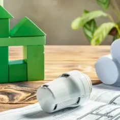 Zielony domek z klocków, termostat i plany budowlane. To wszystko elementy potrzebne do stworzenia energooszczędnego domu z efektywnymi podzielnikami ciepła.