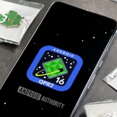 Na ekranie telefonu widnieje logo Androida QPR2 16, sugerujące, jaki jest najnowszy android. Wokół leżą przypinki z zielonym robotem Androida.