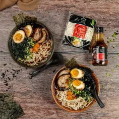 Dwie miski aromatycznego shoyu ramen z makaronem, jajkiem, mięsem i nori. Obok opakowanie makaronu i butelka sosu.