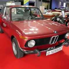Czerwone BMW E10 z lat 70. na czerwonym dywanie, obok motocykle i inne klasyki.