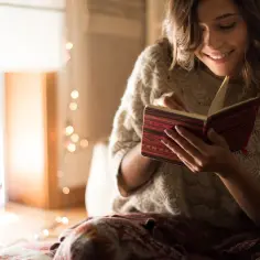 Uśmiechnięta kobieta czyta książkę, szukając w niej cytatów o przemijaniu. W tle migoczą lampki.