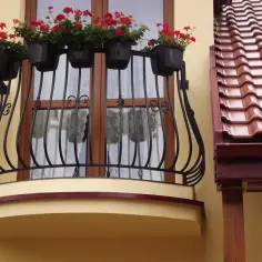 Obróbka czoła balkonu z kwitnącymi pelargoniami. Kute balustrady i drewniane okna dodają uroku.