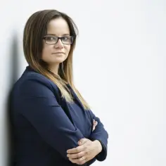 Kobieta w okularach i granatowej marynarce, oparta o ścianę, ze skrzyżowanymi rękami.
