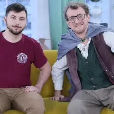 Dwóch mężczyzn, jeden w koszulce, drugi w stroju przypominającym hobbita, siedzi na kanapie.