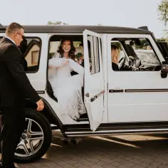 Uśmiechnięta panna młoda w sukni ślubnej wysiada z białego Mercedesa. Zastanawiasz się, ile kosztuje samochód na wesele?