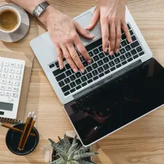 Kobieta pracuje na laptopie, obok kalkulator, kawa i ołówki. Oblicza kwotę wolną od podatku, by zoptymalizować swoje finanse.