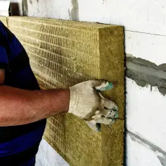 Pracownik w rękawicy montuje płytę izolacyjną do ściany, realizując ocieplenie domu.