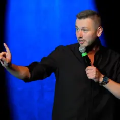 Mężczyzna w czarnej koszuli z mikrofonem, gestykuluje na scenie. To polska stand up.