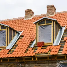 Trwa wymiana okna dachowego. Dach pokryty jest pomarańczową dachówką, a okna są tymczasowo zabezpieczone folią.