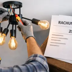 Elektryk w rękawiczkach montuje żyrandol, obok leży rachunek na 250 zł.