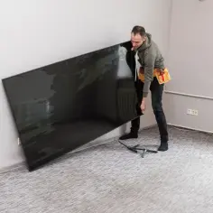 Mężczyzna z pasem narzędziowym przy ścianie trzyma duży, czarny telewizor, zastanawiając się, na jakiej wysokości telewizor będzie najlepiej wyglądał.