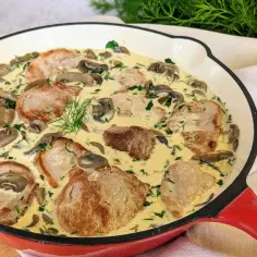Pyszne polędwiczki wieprzowe w kremowym sosie z pieczarkami i koperkiem, podane w czerwonej patelni.