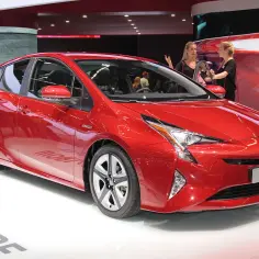 Błyszczący, czerwony Prius 4 na wystawie. Dwie kobiety prezentują samochód, a w tle widać innych zwiedzających.