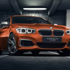 Pomarańczowe BMW 140i z tabliczką "SUPERCAR CLUB" stoi w garażu.