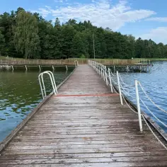Drewniany pomost prowadzi do spokojnego jeziora Luterskiego, otoczonego zielonym lasem.