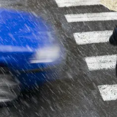 Deszczowa pogoda na przejściu dla pieszych. Niebieski samochód mija kobietę z parasolem.