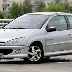 Srebrny Peugeot 206 S16 z napisem "Quiksilver" na błotniku, zaparkowany na mokrym parkingu.