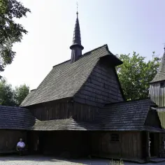 Drewniany kościół z wieżą, otoczony drzewami. Na ławce przed kościołem siedzi starszy pan, jakby opowiadał historie o dawnych czasach, gdy takie **cmolas** były powszechne.
