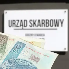 Złotówki przed tablicą "Urząd Skarbowy". Zastanawiasz się, ile czasu na zapłacenie mandatu? Sprawdź godziny otwarcia urzędu.
