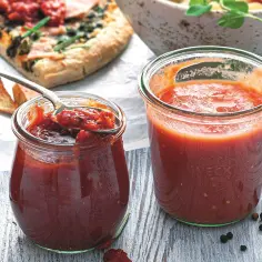 Dwa słoiki z domowym sosem pomidorowym, idealnym do pizzy. Jeden otwarty, z łyżką pełną sosu, obok kawałek pizzy.