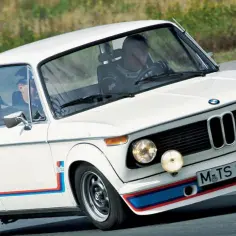 Białe BMW 2002 Turbo z kolorowymi pasami na błotnikach i zderzaku, pędzi po torze.