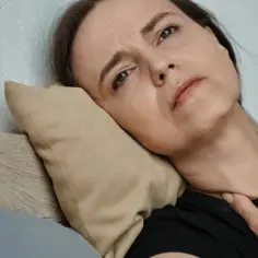 Kobieta leży na poduszce, trzymając się za szyję, z wyrazem bólu i duszności na twarzy.
