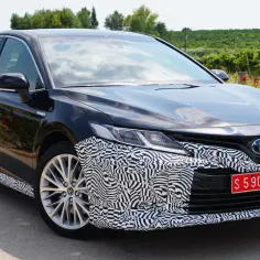 Czarny prototypowy Toyota Camry kombi w kamuflażu, zaparkowany na tle winnicy i kwitnących róż.