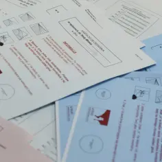 Karta do głosowania w wyborach samorządowych. Na stole leżą pakiety kart do głosowania, niektóre białe, inne niebieskie i różowe.