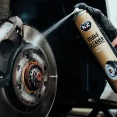 Czyszczenie hamulców z użyciem preparatu K2 Brake Cleaner. Tarcza hamulcowa wygląda na przegrzaną, pokryta rdzą i brudem.