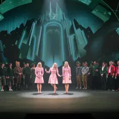Trzy dziewczyny w różowych sukienkach na scenie, w tle zielony pałac Oz. To scena z musicalu "Wicked", który ma imponujący czas trwania.